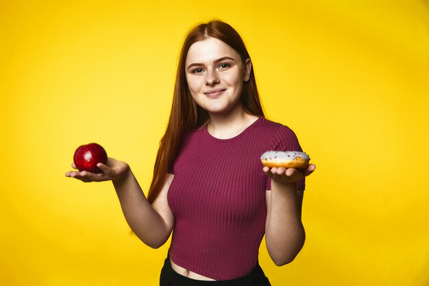 Smiled redhead caucasian girl is holding apple in one hand and donut in another hand