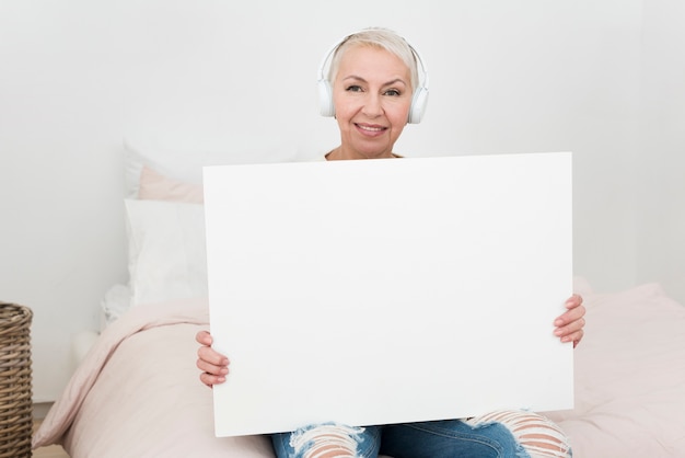 Smiley elderly woman wearing headphones and holding blank placard in bed