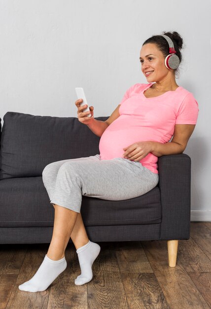 Smiley pregnant woman with smartphone listening to music on headphones at home