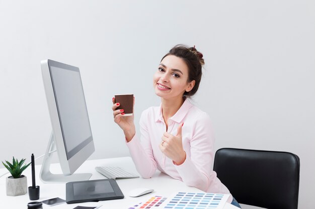 Smiley woman at desk holding cup of coffee and giving thumbs up