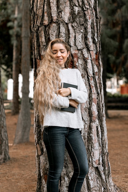 Free Photo smiley woman standing next to a tree while holding a book