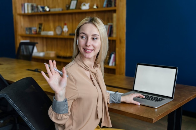 Free photo smiley woman waving in the office while working on laptop