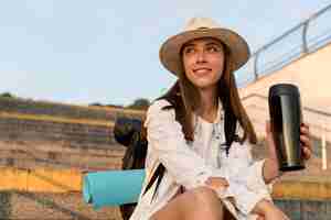 Free photo smiley woman with backpack and hat holding thermos while traveling