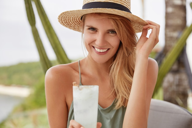 Smiling adorable woman with happy expression has summer vacation, spends free time in outdoor cafe with fresh cold drink, looks positively. Attractive female in straw hat being in good mood.