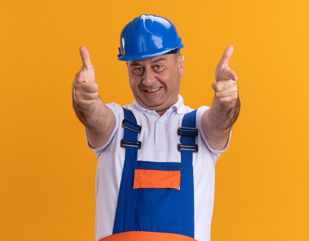 Smiling adult builder man in uniform points at front with two hands isolated on orange wall