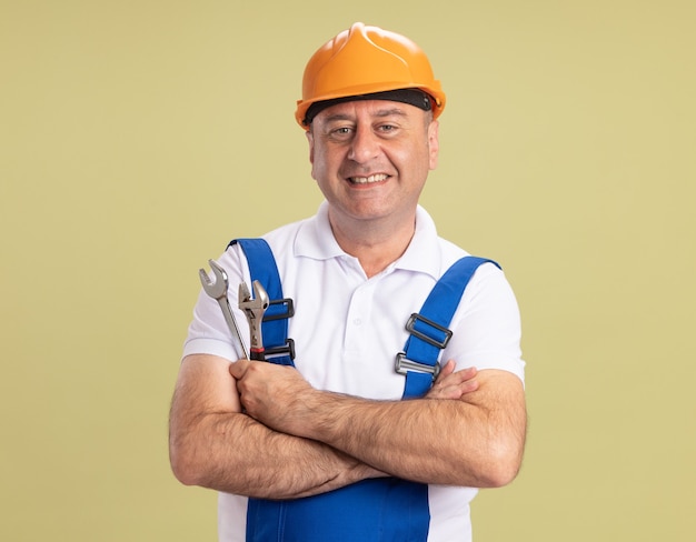 Free Photo smiling adult builder man in uniform stands with crossed arms holding wrench and monkey wrench isolated on olive green wall