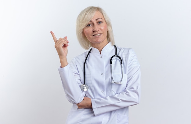 Free photo smiling adult slavic female doctor in medical robe with stethoscope pointing up isolated on white background with copy space