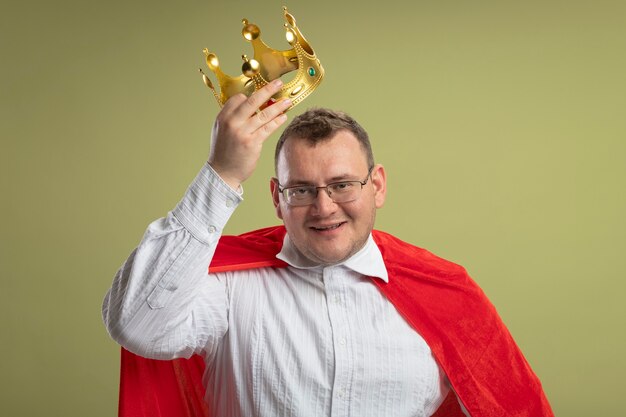 Smiling adult slavic superhero man in red cape wearing glasses holding crown above head  isolated on olive green wall