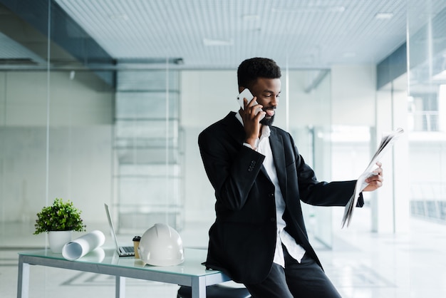 Smiling african american businessman talking on the phone with newspaper and cup of coffee in office