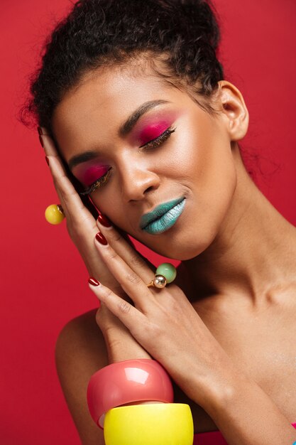 smiling african american woman with bright colorful makeup putting head on palm, isolated over red wall