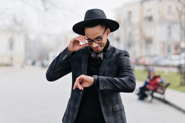 Free photo smiling african guy looking at wristwatch while standing on the street. outdoor portrait of happy young man waiting girlfriend in park.