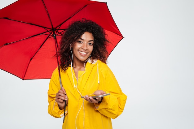 Smiling african woman in raincoat hiding under umbrella and listening music