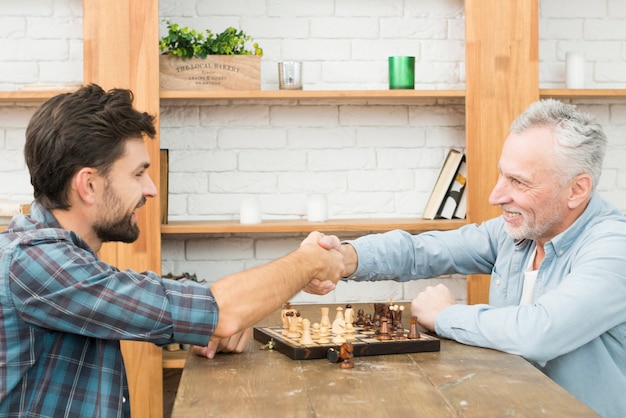 Smiling aged man and young guy shaking hands at table with chess board