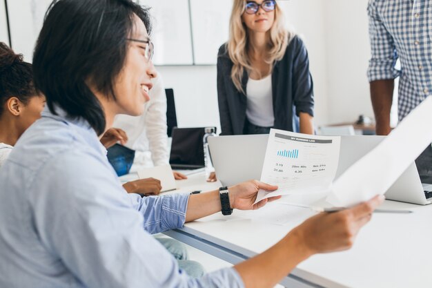 Smiling asian businessman analysing infographic in his office. Indoor portrait of young it-specialists with chinese developer in glasses