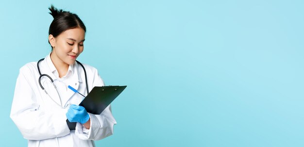 Free Photo smiling asian doctor nurse writing down on clipboard standing in medical uniform over blue backgroun