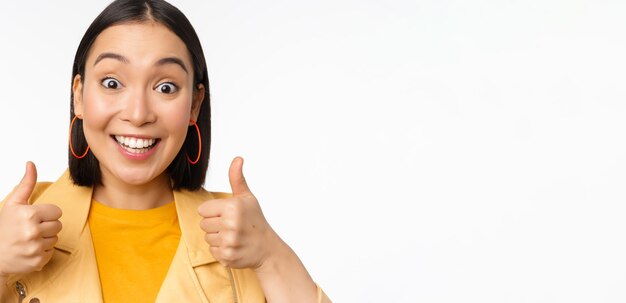 Smiling asian girl showing thumbs up looking pleased approve smth standing over white background