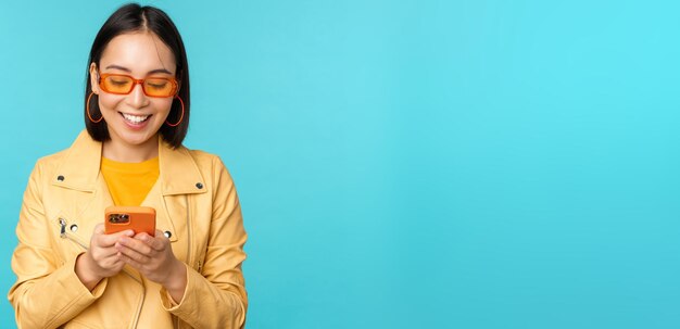 Smiling asian girl in sunglasses using smartphone app holding mobile phone standing over blue backgr