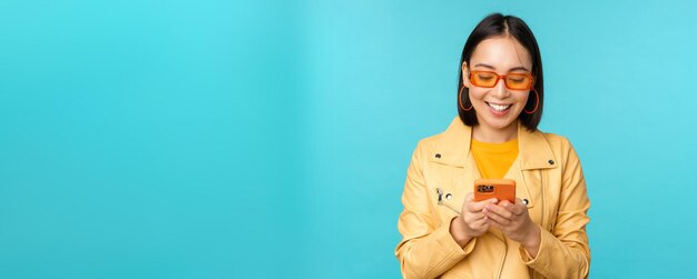 Smiling asian girl in sunglasses using smartphone app holding mobile phone standing over blue backgr