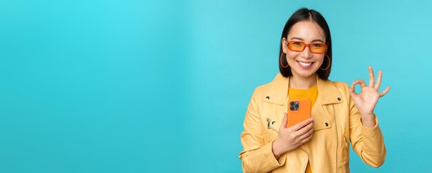 Smiling asian girl with smartphone shwoing okay ok sign in approval standing over blue background Copy space