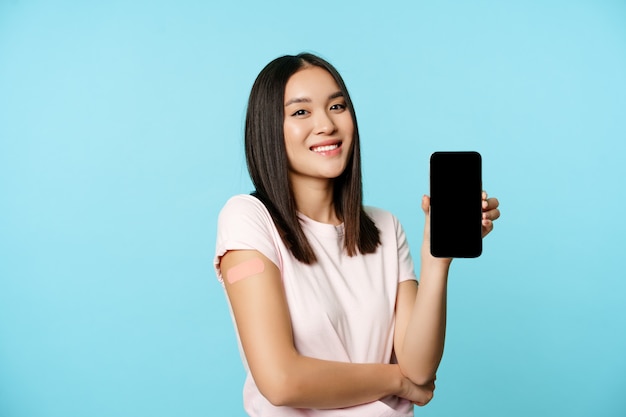 Free photo smiling asian girl with vaccinated arm, showing mobile phone empty screen, concept of health passport, covid-19 vaccination certificate on smartphone