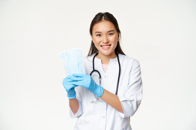 Smiling asian woman doctor, nurse showing medical face masks, wearing sterile rubber gloves, standing over white background