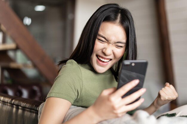 Smiling asian woman looking satisfied making fist pump and reading mobile phone sitting at home and ...