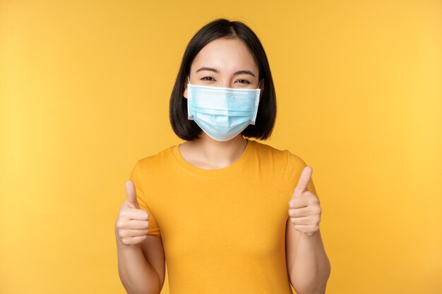 Smiling asian woman in medical face mask showing thumbs up approval like and recommend smth standing over yellow background