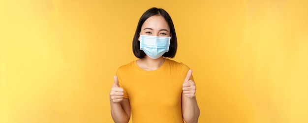 Smiling asian woman in medical face mask showing thumbs up approval like and recommend smth standing over yellow background