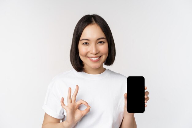 Smiling asian woman showing okay mobile phone screen recommending smartphone app standing over white background