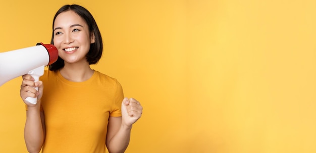 Smiling asian woman standing with megaphone announcing smth advertising product standing over yellow