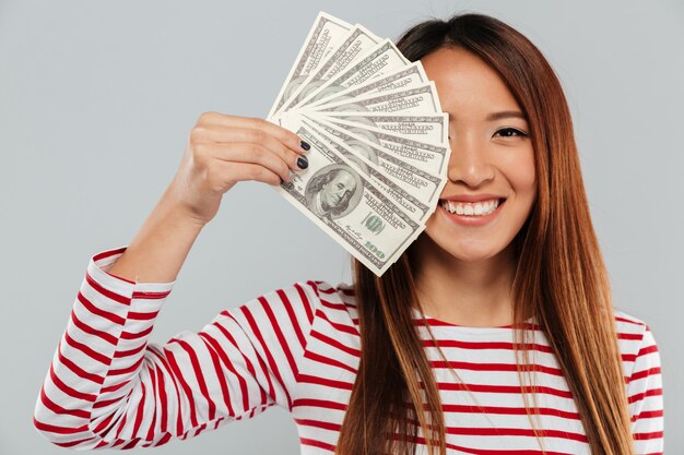 Smiling asian woman in sweater is covering in money of half of face and looking at the camera over gray backgound