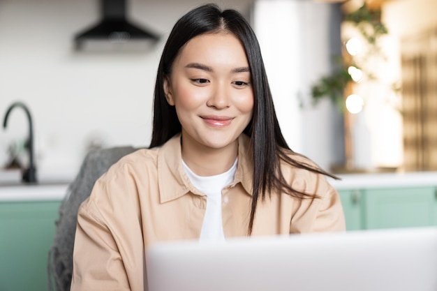 Smiling asian woman using laptop from home girl sits in kitchen with computer distance education con...