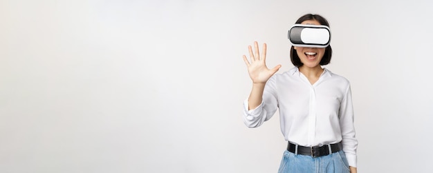 Free Photo smiling asian woman waving at you wearing virtual reality glasses vr headset chatting in meta universe standing over white background