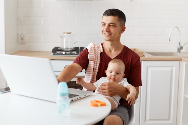 Smiling attractive brunette man freelancer wearing casual style maroon t shirt, working online and looking after his little baby daughter, using laptop for working.