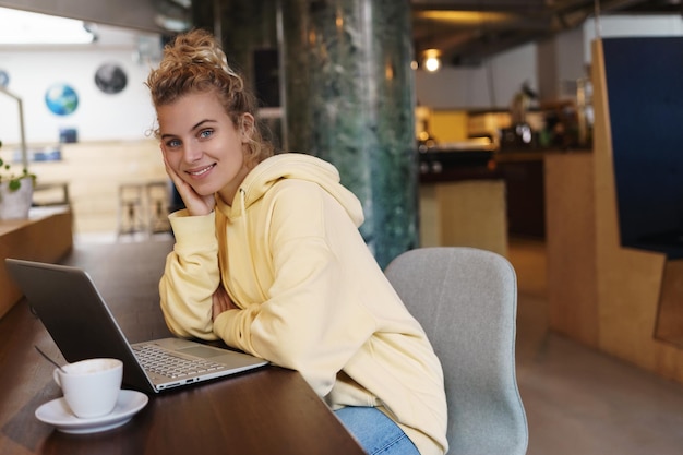 Free photo smiling attractive woman sitting in cafe drinking coffee and using laptop beautiful girl looking at camera happy while working remote from cafe female student studying outdoors using laptop