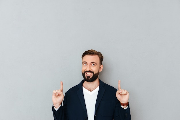 Smiling bearded man in busines clothes pointing and looking up over gray 