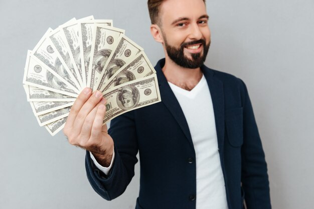 Smiling bearded man in busines clothes showing money and looking at the camera over gray 