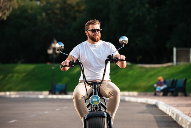 Free photo smiling bearded man in sunglasses rides on modern motorbike outdoors and looking away