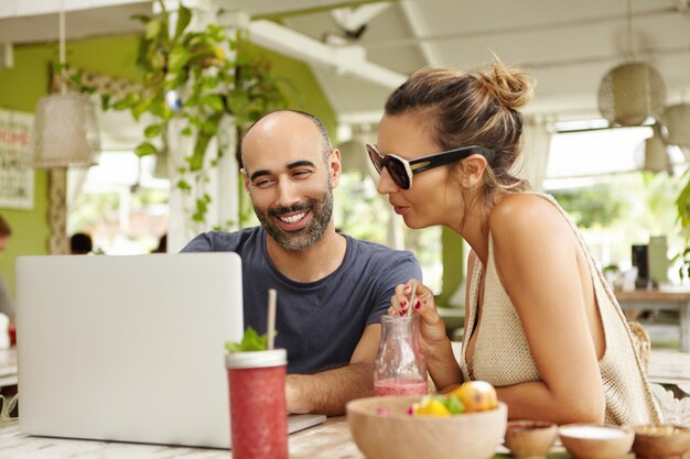 Smiling bearded man and woman in sunglasses sitting in front of open laptop and discussing something, looking at screen with interest.