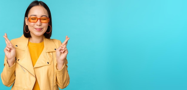 Smiling beautiful asian woman wishing cross fingers for good luck and looking hopeful standing over blue background