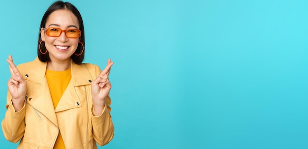 Smiling beautiful asian woman wishing cross fingers for good luck and looking hopeful standing over