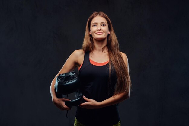 Smiling beautiful brunette female boxer in sportswear holds a protective helmet, looking at a camera. Isolated on a dark textured background.