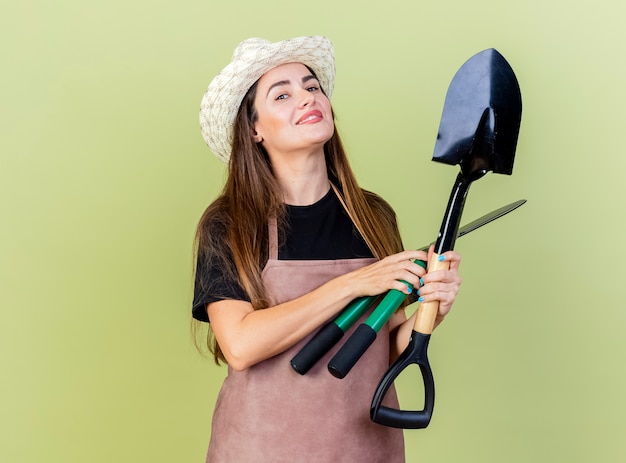 Smiling beautiful gardener girl in uniform wearing gardening hat holding clippers with spade isolated on olive green background