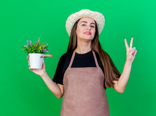 Smiling beautiful gardener girl in uniform wearing gardening hat holding flower in flowerpot showing peace gesture isolated on green background