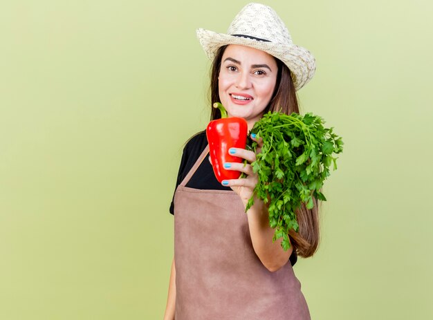 Smiling beautiful gardener girl in uniform wearing gardening hat holding out cliantro with pepper at camera isolated on olive green background