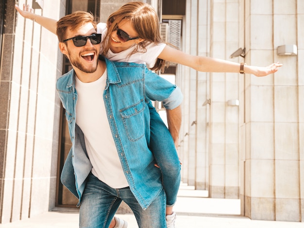 Free photo smiling beautiful girl and her handsome boyfriend in casual summer clothes. man carrying his girlfriend on the back and she raising her hands.