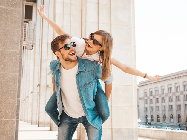 Smiling beautiful girl and her handsome boyfriend in casual summer clothes. Man carrying his girlfriend on the back and she raising her hands. 