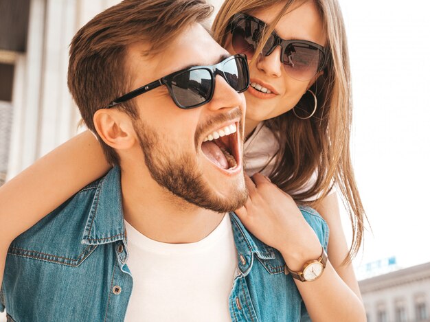 Smiling beautiful girl and her handsome boyfriend in casual summer clothes. Man carrying his girlfriend on the back and she raising her hands. 