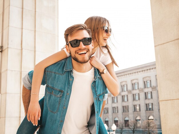 Smiling beautiful girl and her handsome boyfriend in casual summer clothes. Man carrying his girlfriend on the back and she raising her hands. 