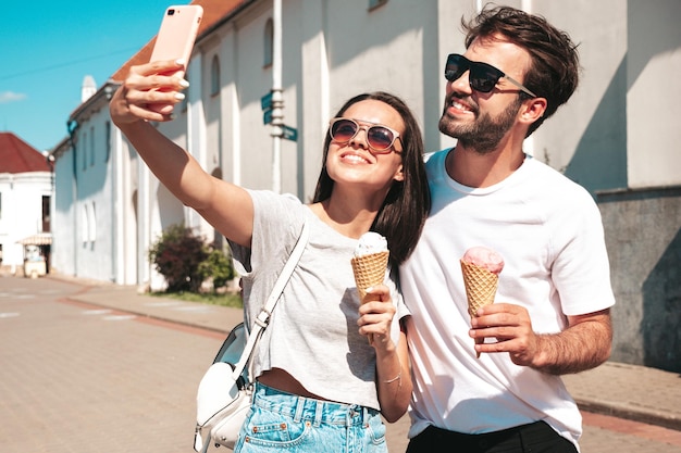 Smiling beautiful woman and her handsome boyfriend Woman in casual summer clothes Happy cheerful family Couple posing in the street Eating tasty ice cream in waffles cone Taking selfie photos